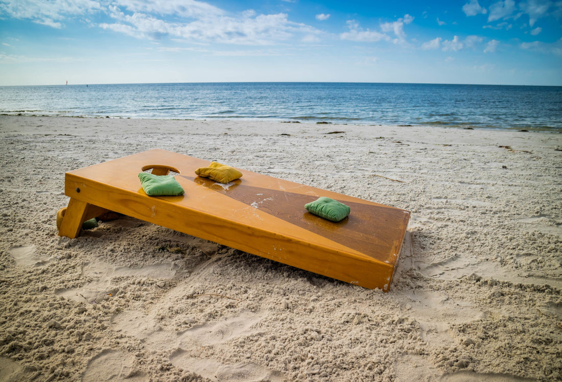 Bean bag toss board on a sandy beach overlooking the water and blue sky, with bean bag toss bags filled with Victory Pellets resting on top–durable, moisture-resistant, and made to last.