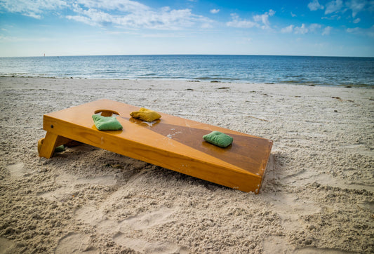 Bean bag toss board on a sandy beach overlooking the water and blue sky, with bean bag toss bags filled with Victory Pellets resting on top–durable, moisture-resistant, and made to last.